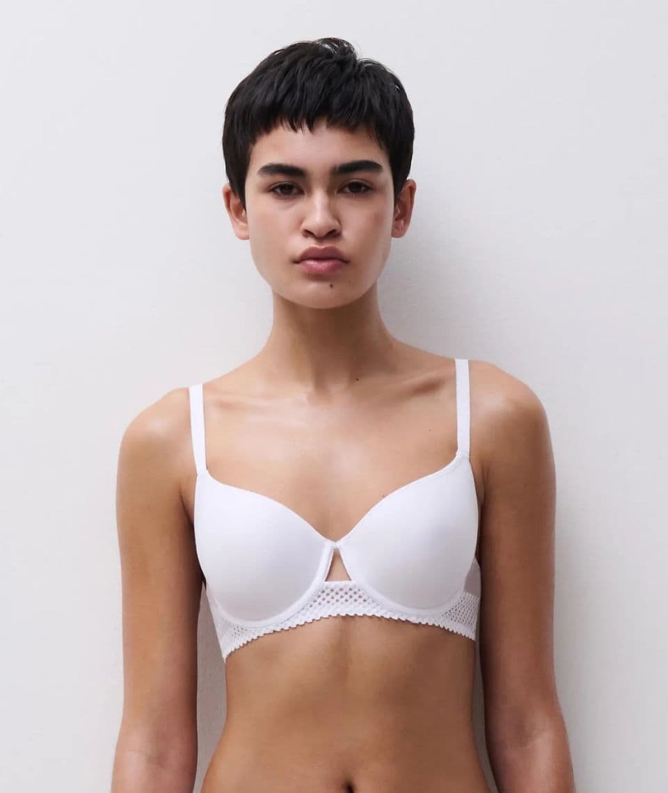 Person with short dark hair wearing a white bra, standing against a plain light background.