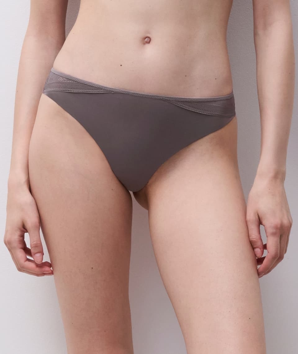 Person wearing taupe bikini briefs with subtle lace detail on the sides, standing against a neutral background.