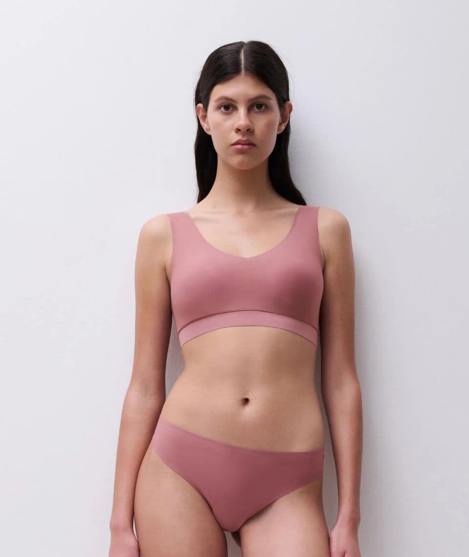 Woman in a pink underwear and bra stands against a white background, with her hair down, looking directly at the camera.