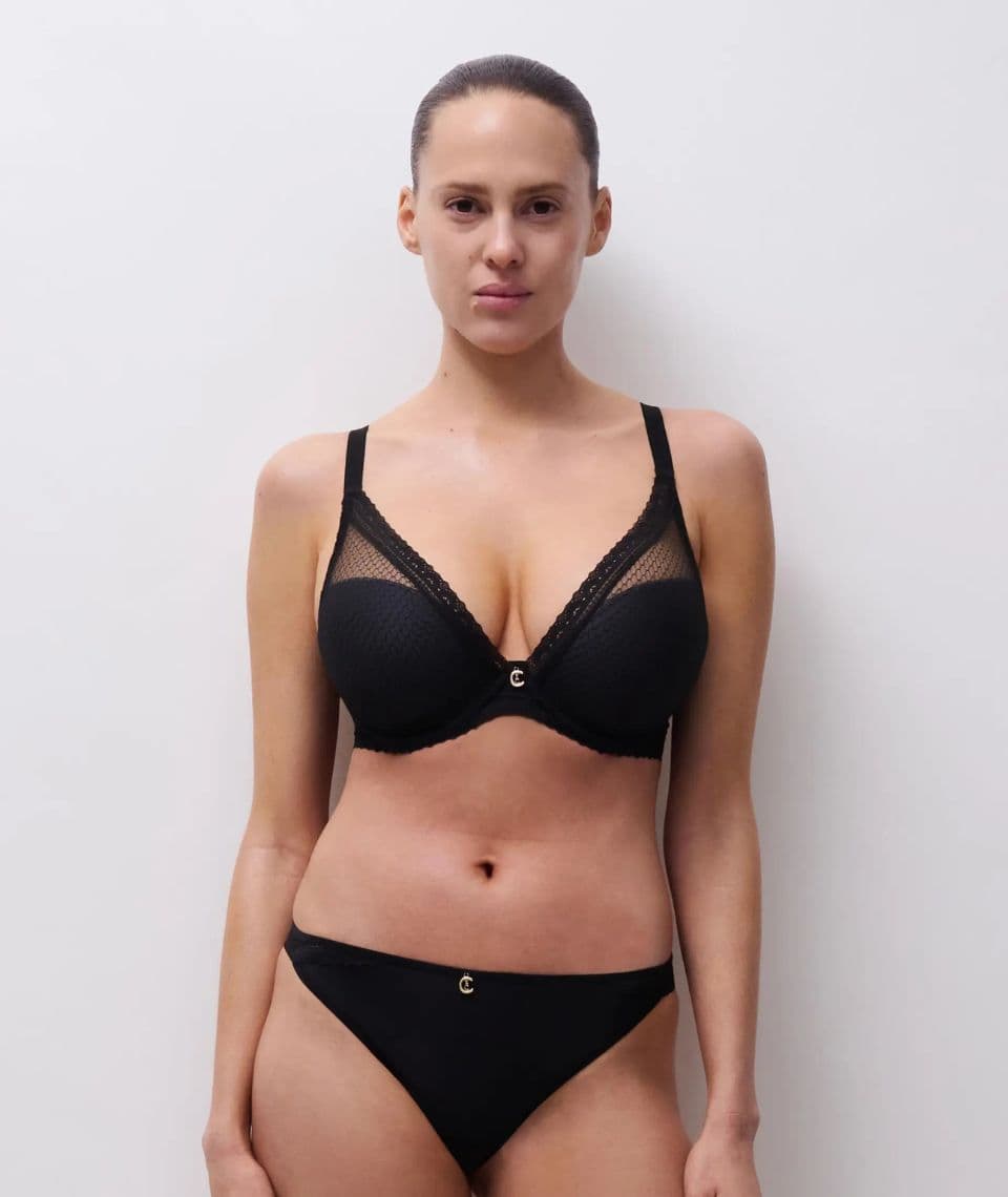 A woman in a black bra and underwear stands against a plain white background, facing forward with a neutral expression.