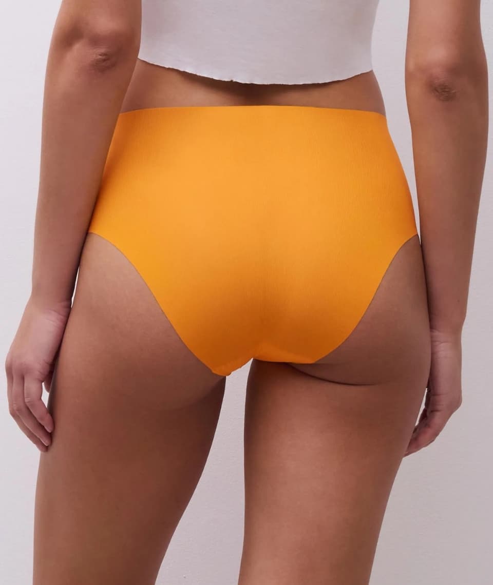 Back view of a person wearing high-waisted orange underwear and a white cropped top, standing against a light background.