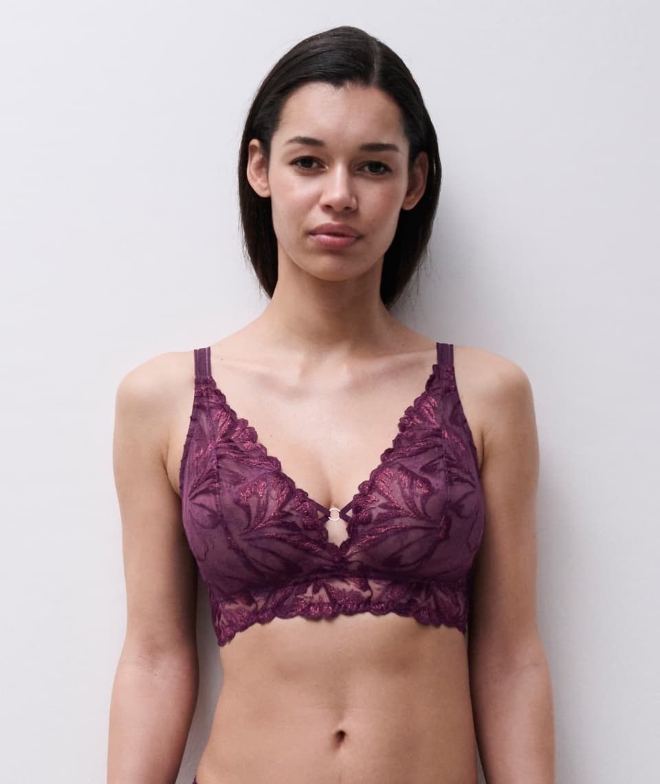 A woman wearing a dark lace bralette with floral designs, standing against a plain light gray background.