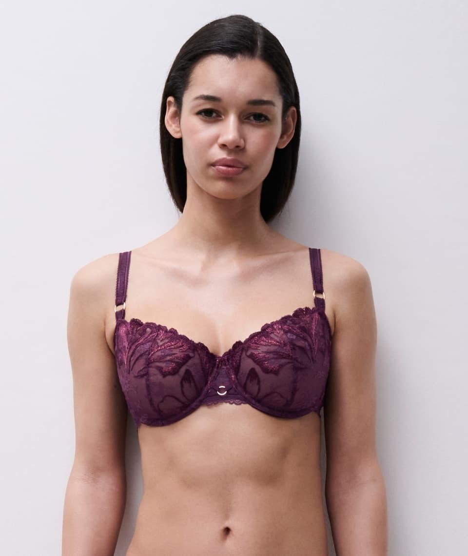 Person wearing a dark lace bra with floral patterns, standing against a light gray background.