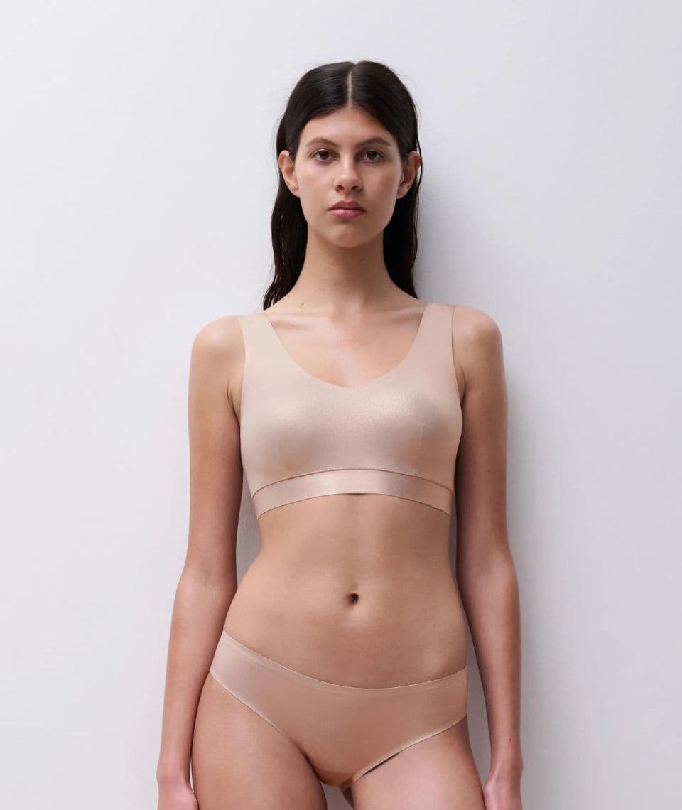 Person with long dark hair in light undergarments stands against a plain white background, looking forward.