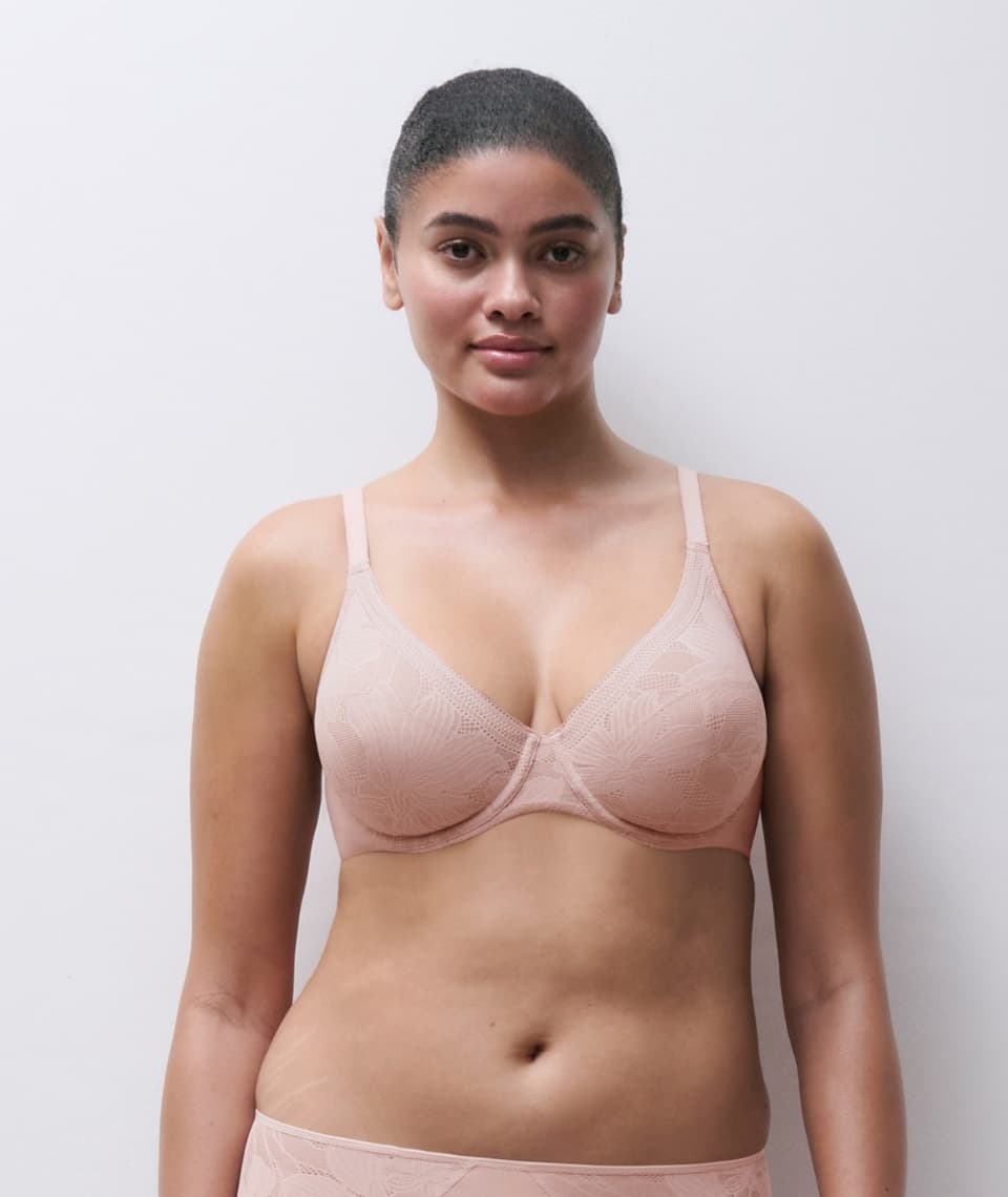 A woman wearing a light pink, lace-patterned bra and matching underwear, standing against a plain white background.