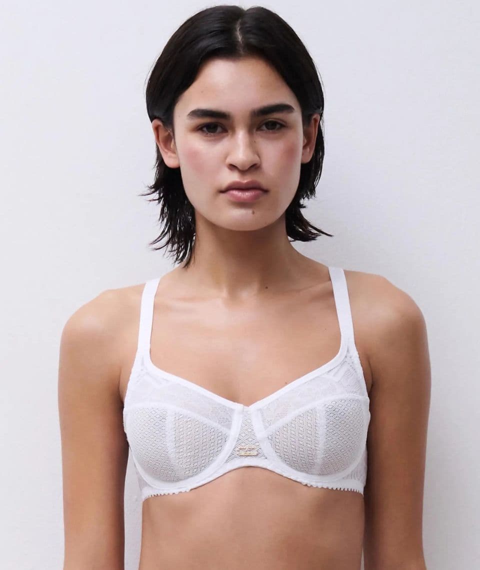 A person wearing a white lace bra, standing against a plain light background, looking directly at the camera.