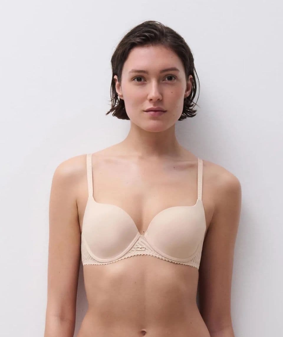 Person with short hair wearing a beige bra, standing against a plain white background.