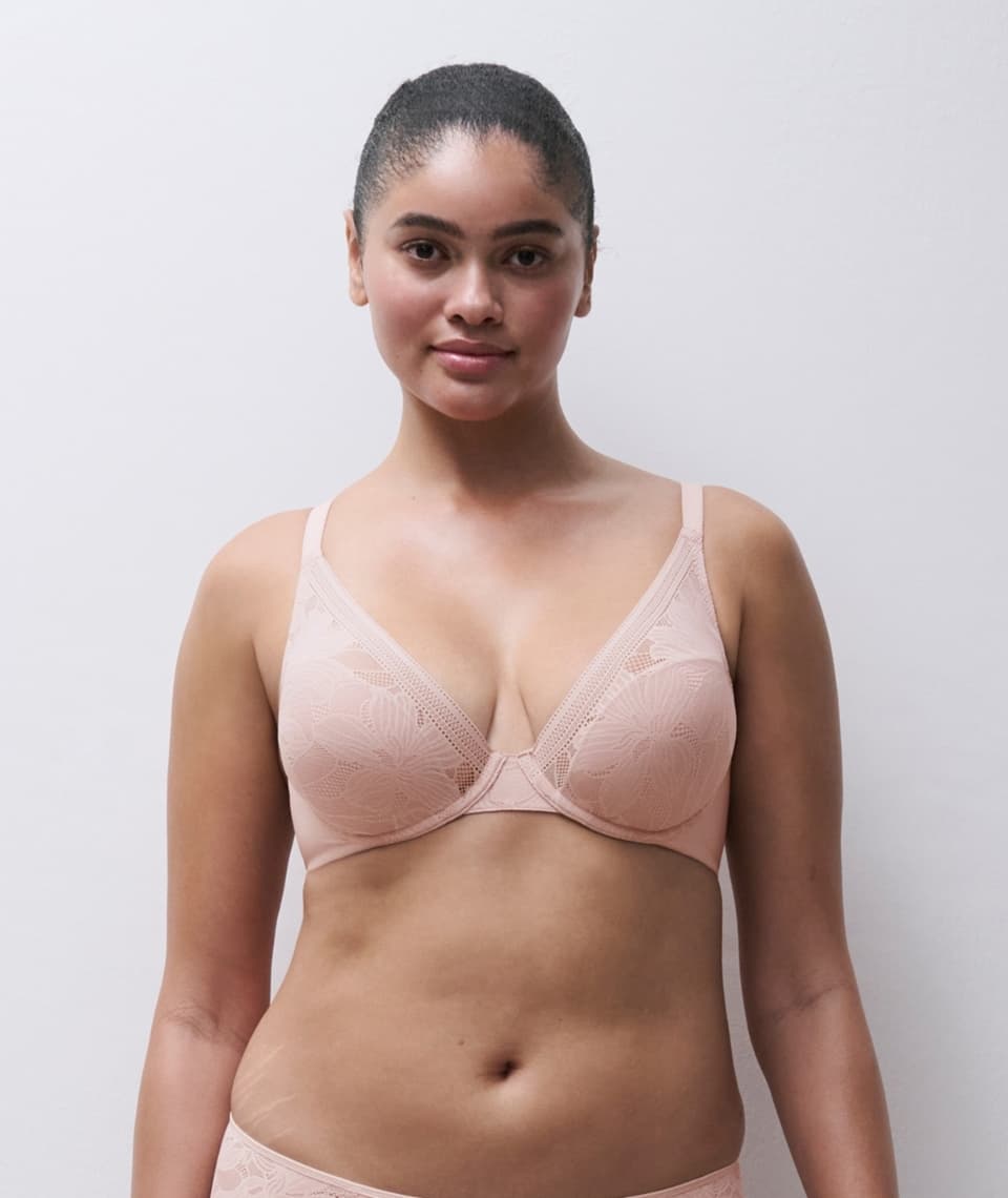 A woman wearing a light pink lace bra stands against a plain white background, looking confidently into the camera.