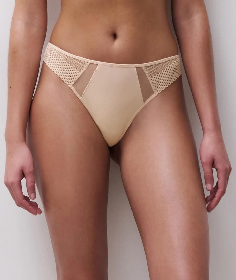Woman in beige lace-trimmed panties, with mesh panels, standing against a plain background.