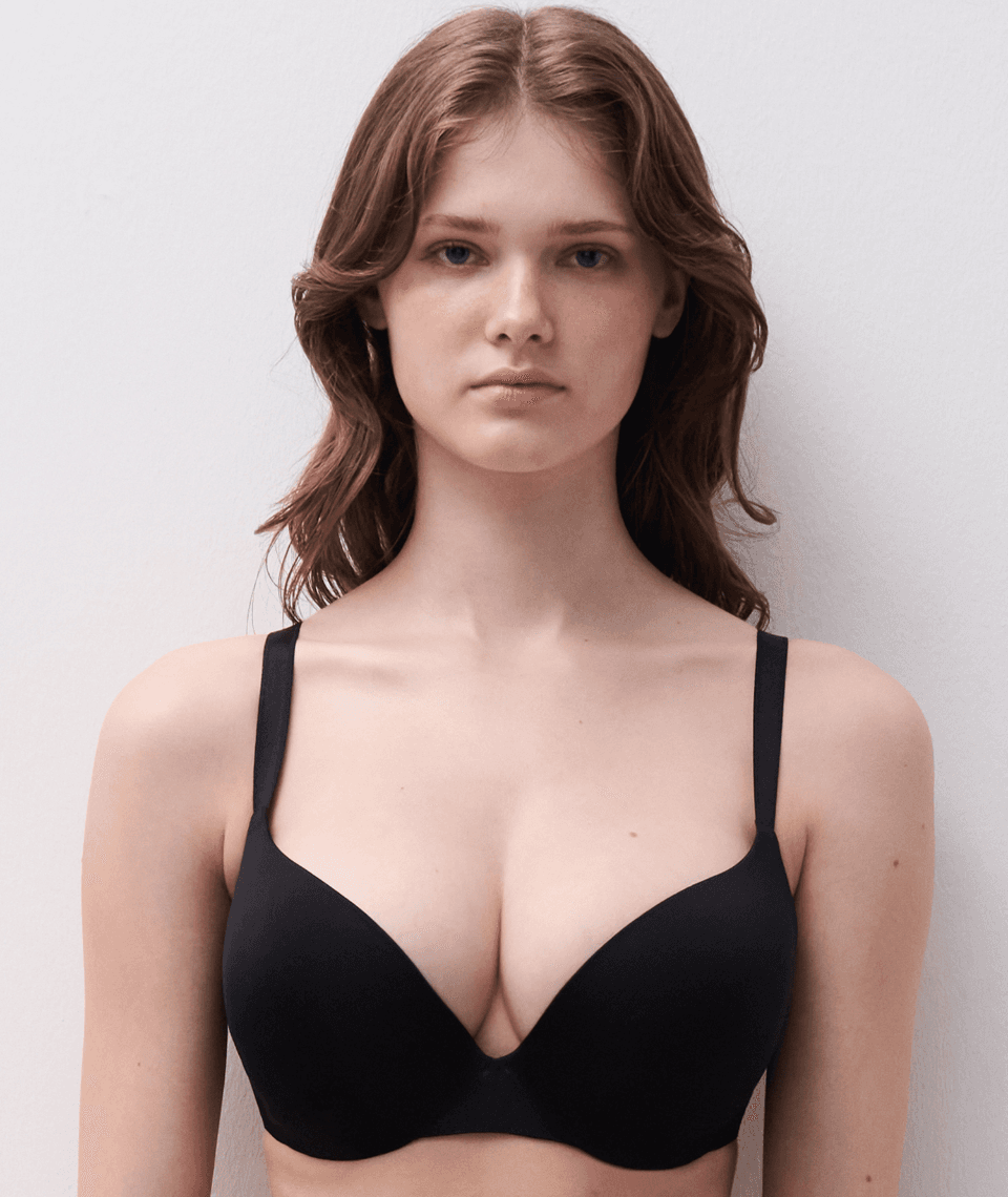 A person with long brown hair wears a black bra, standing against a plain white background, looking directly at the camera.