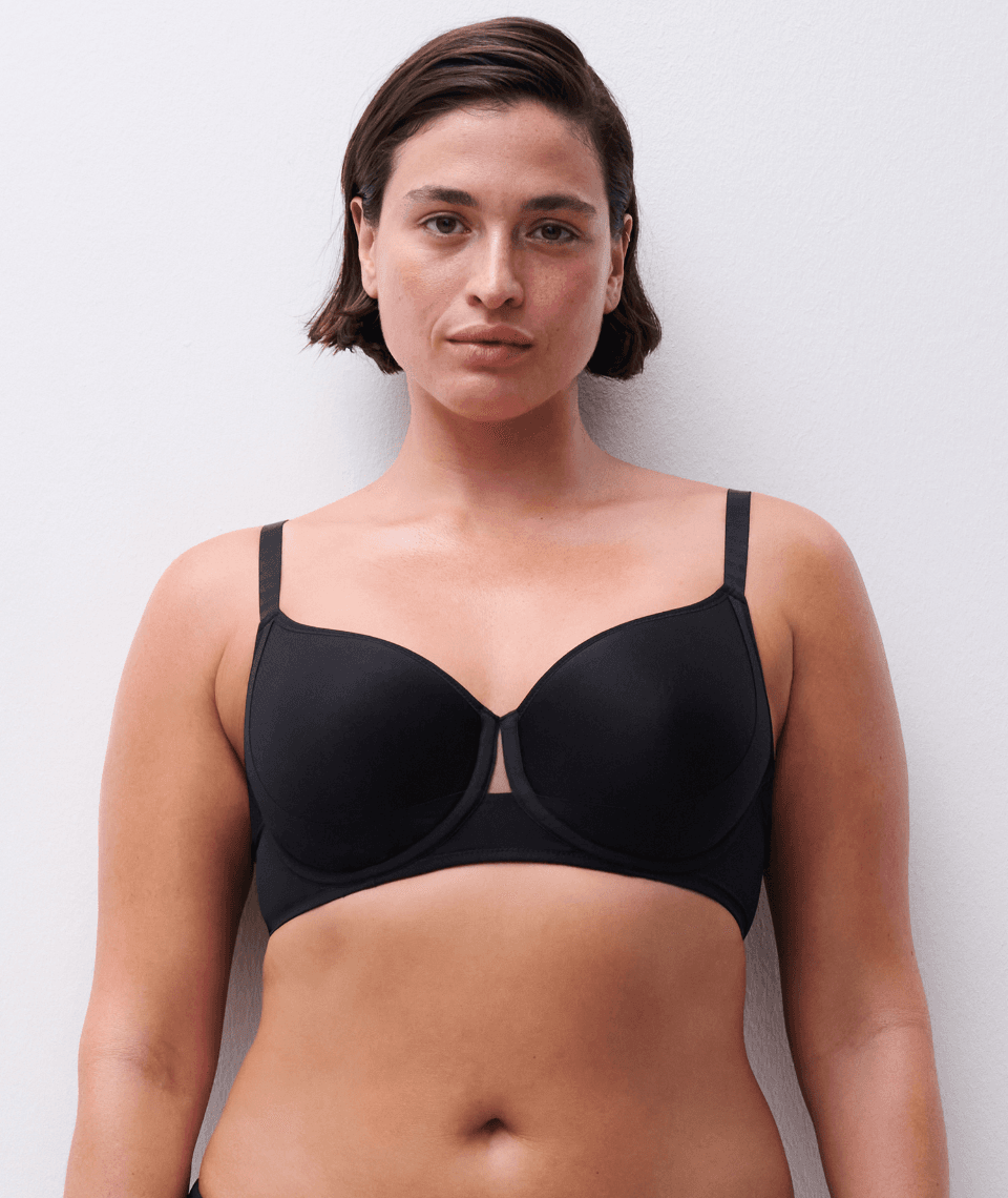 A woman with short dark hair wears a black bra, standing against a plain white background.