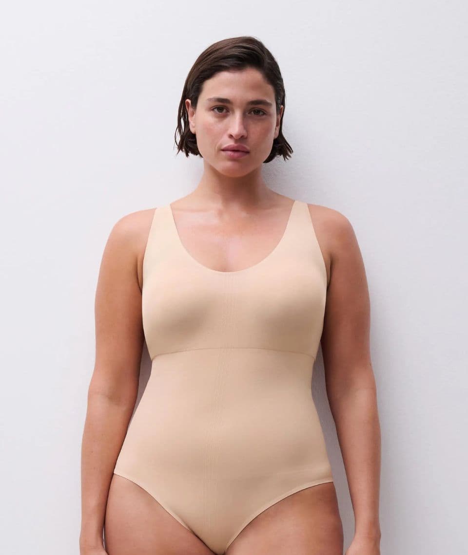 A person wearing a nude shapewear body standing against a plain white background, looking directly at the camera.