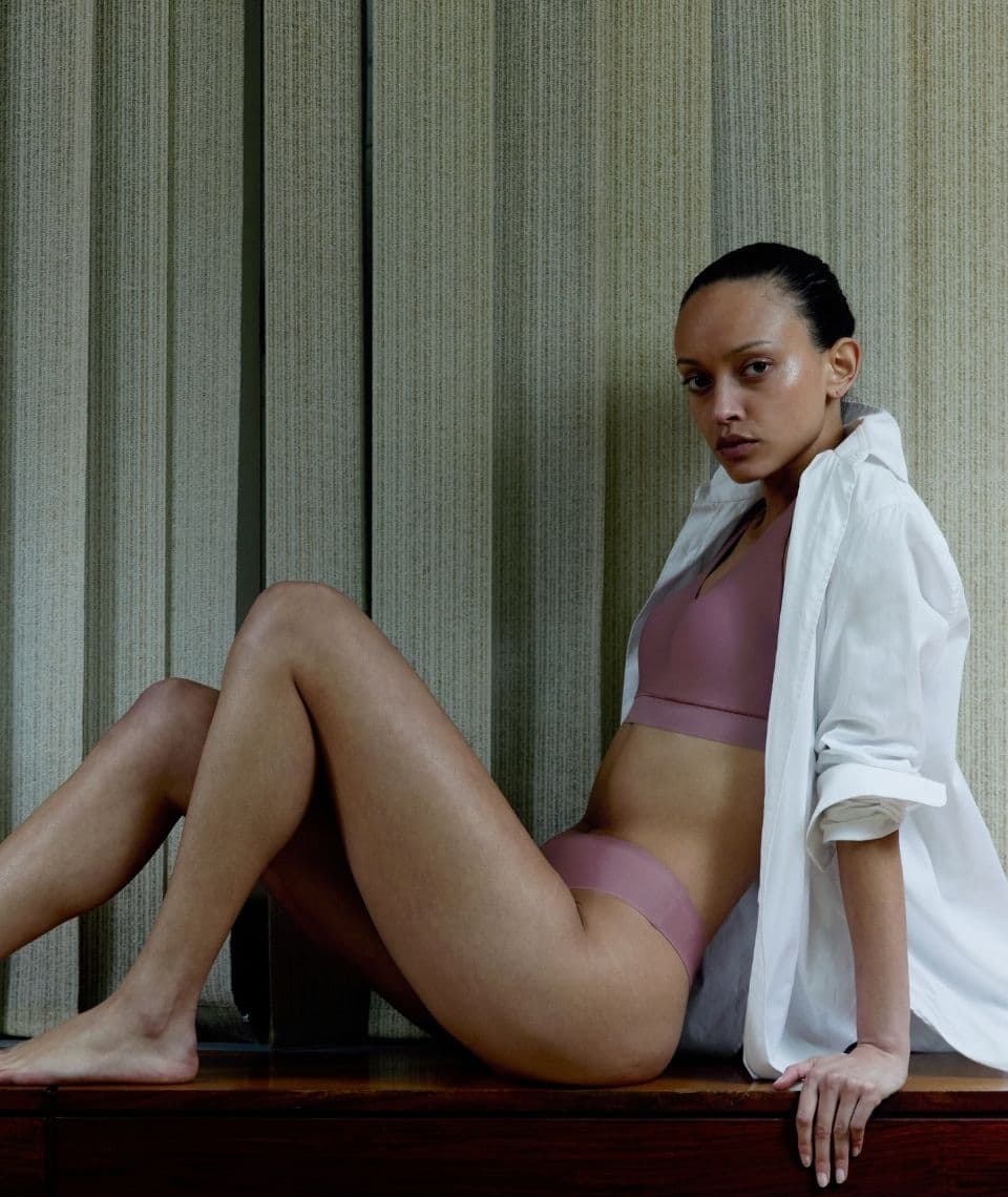 Person in mauve underwear and a white shirt sits on a wooden surface against vertical blinds, looking intently at the camera.