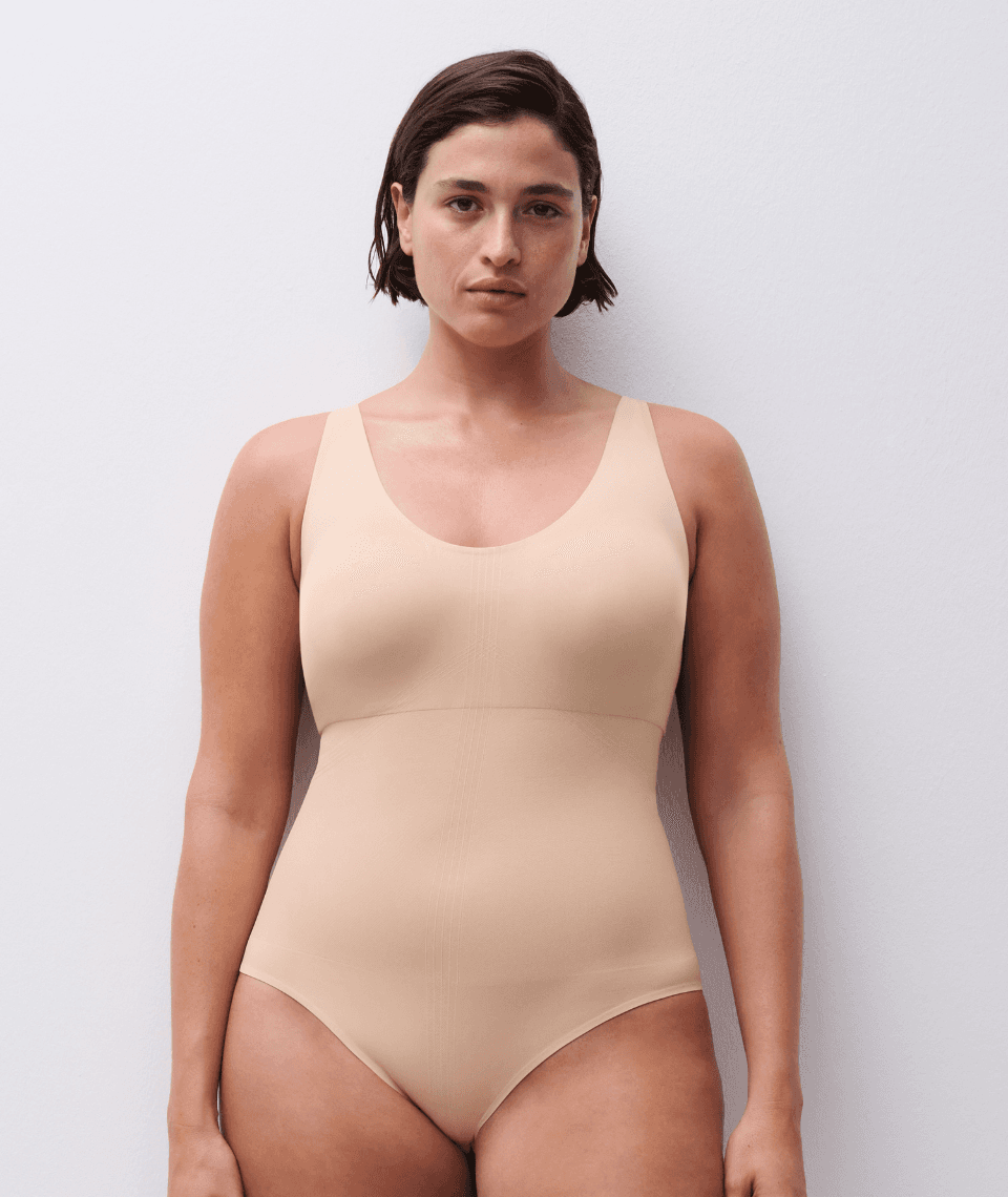 A person wearing a nude seamless bodysuit stands against a plain white background, looking directly at the camera.