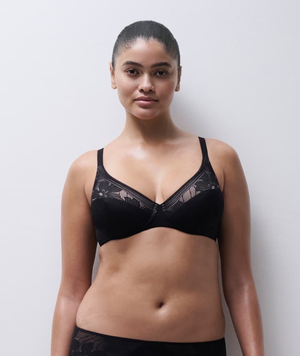 Person modeling a black lace bra with floral patterns, standing against a plain white background.