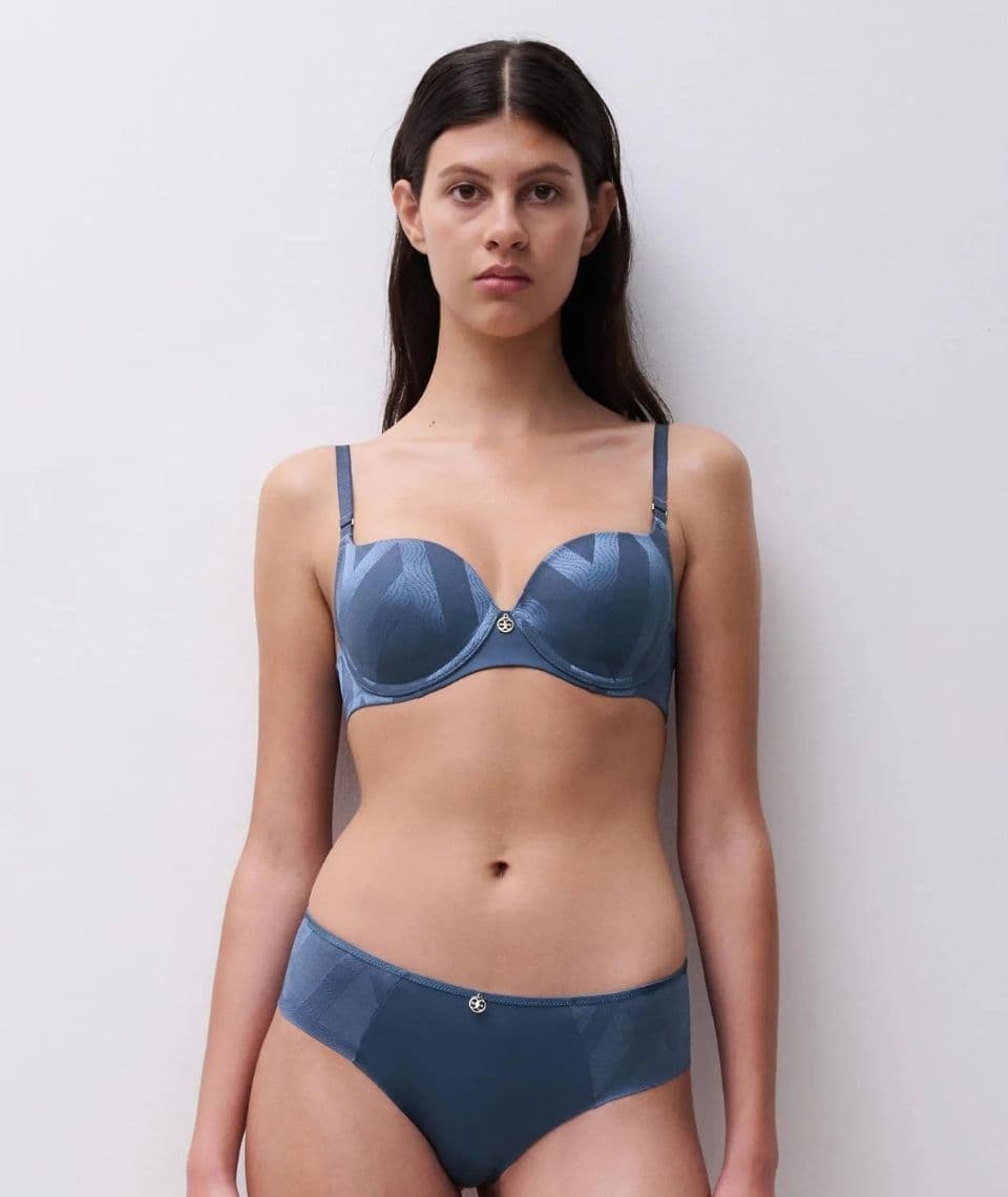 A woman stands against a plain background, wearing a blue lingerie set. She looks directly at the camera, with long dark hair and a neutral expression.