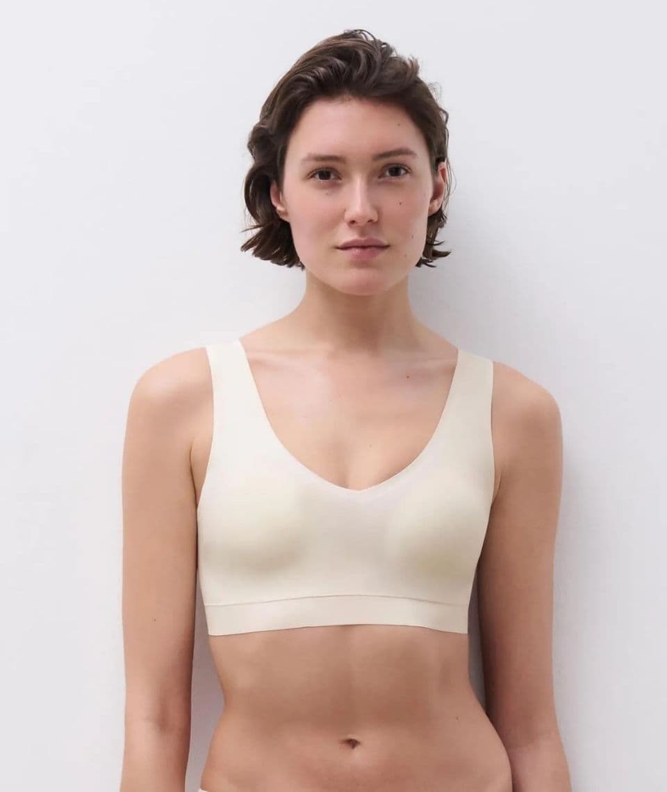 A person with short brown hair wears a white bra, standing against a plain white background, looking directly at the camera.