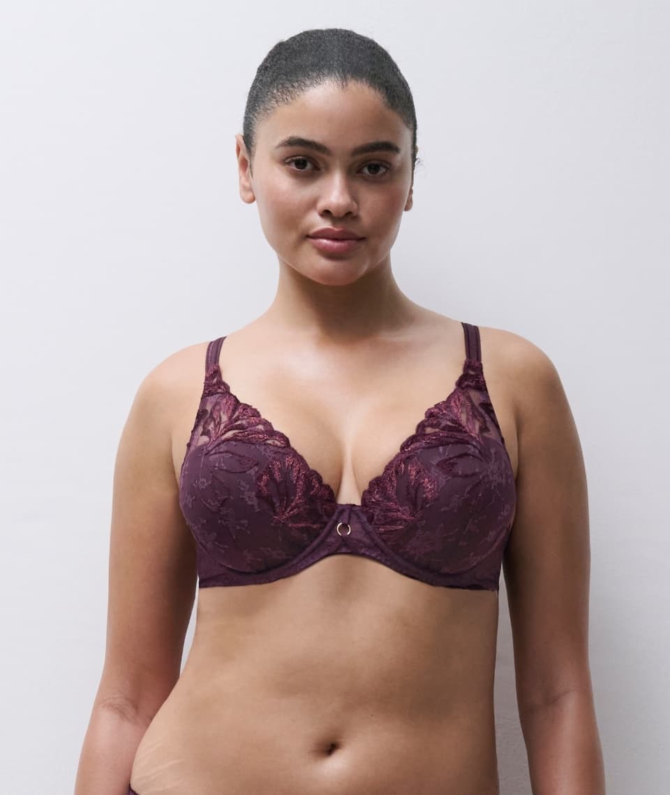 A woman models a dark lace bra with floral embroidery, standing against a plain white background.