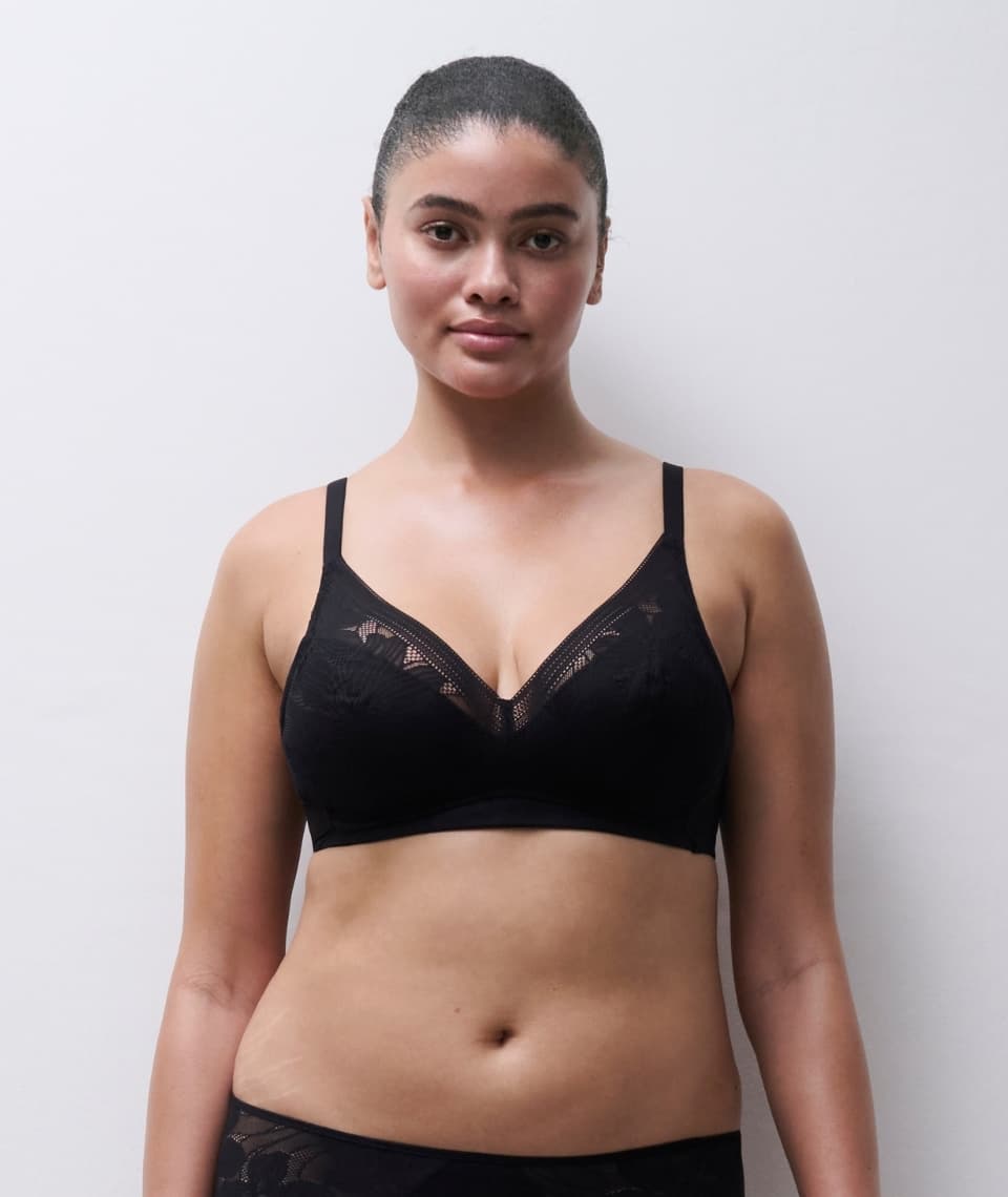 Person wearing a black lace bra, standing against a white background, looking confidently at the camera.