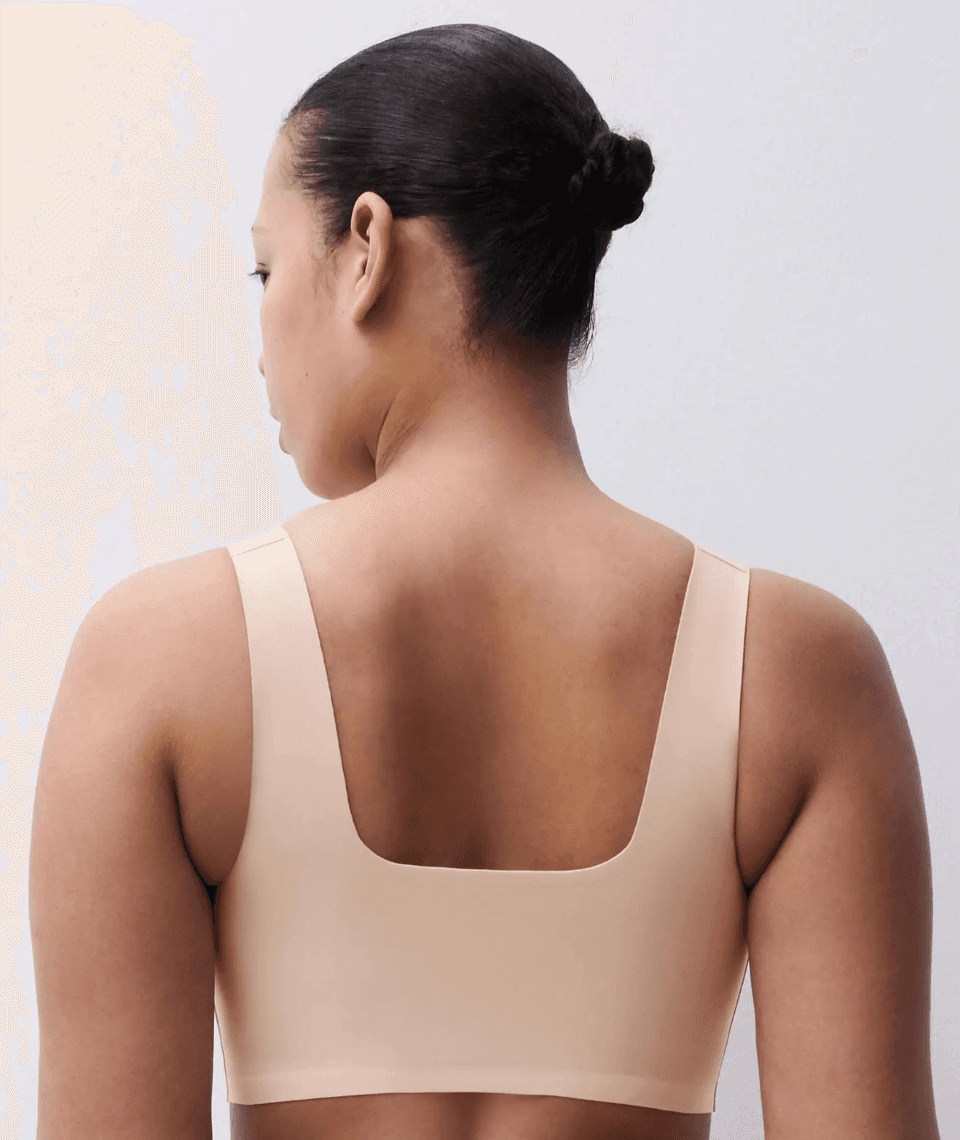 Person with hair in a bun wearing a beige bra, facing away from the camera against a plain background.