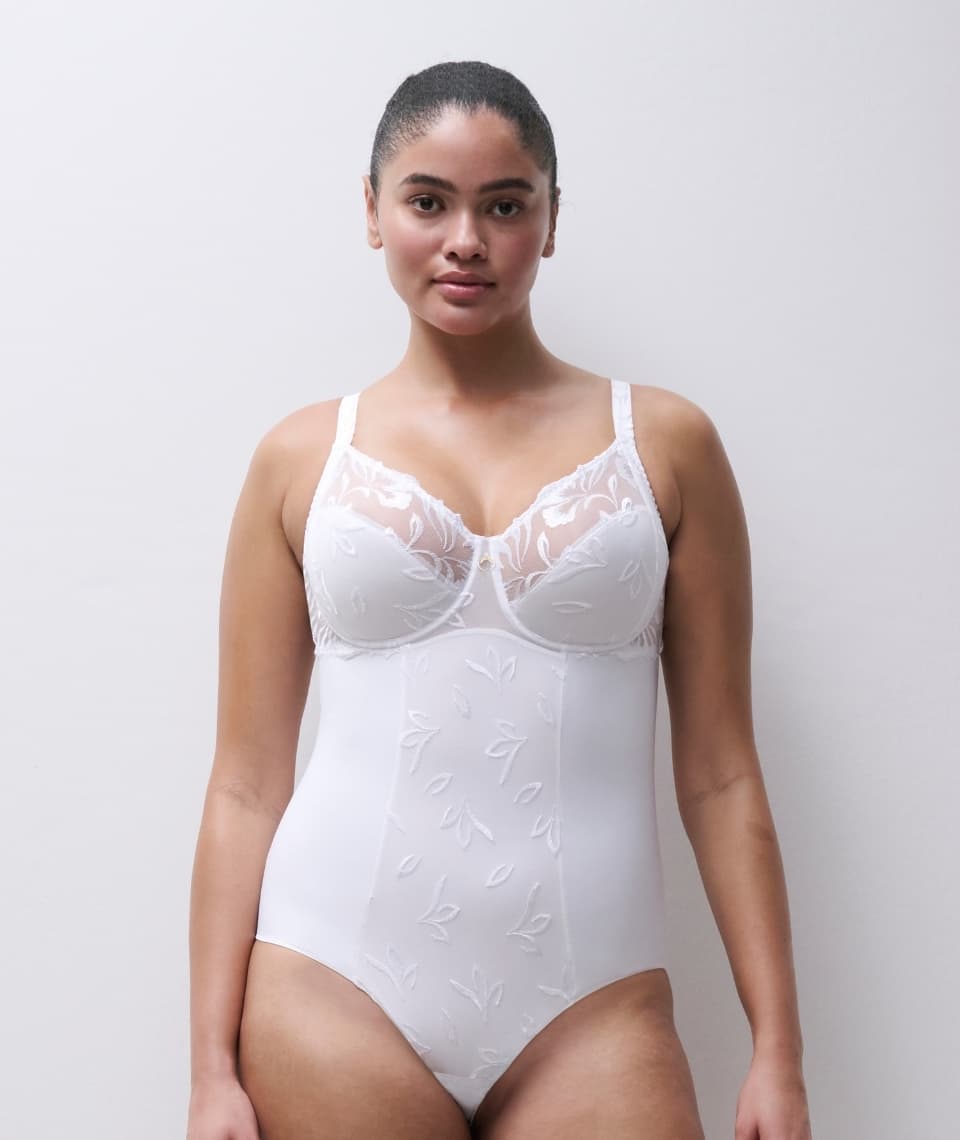 A person wearing a white lace bodysuit stands against a plain background, showcasing the garment's delicate floral patterns.
