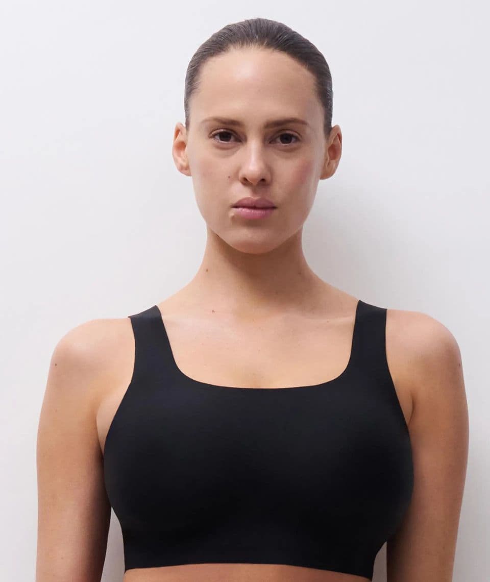Person with tied-back hair wearing a black bra against a plain background, looking directly at the camera.