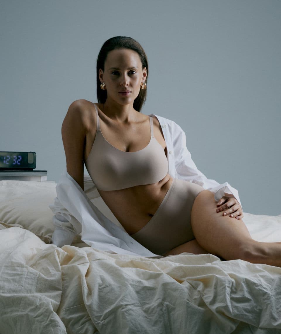A person in beige underwear and an open white shirt sits on a bed beside a digital clock displaying 12:32.