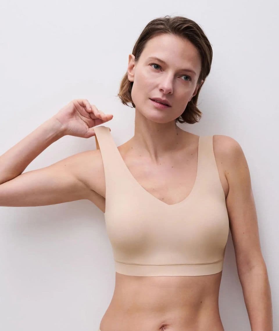 A woman in a beige bra adjusts the strap while looking off-camera, standing against a plain white background.
