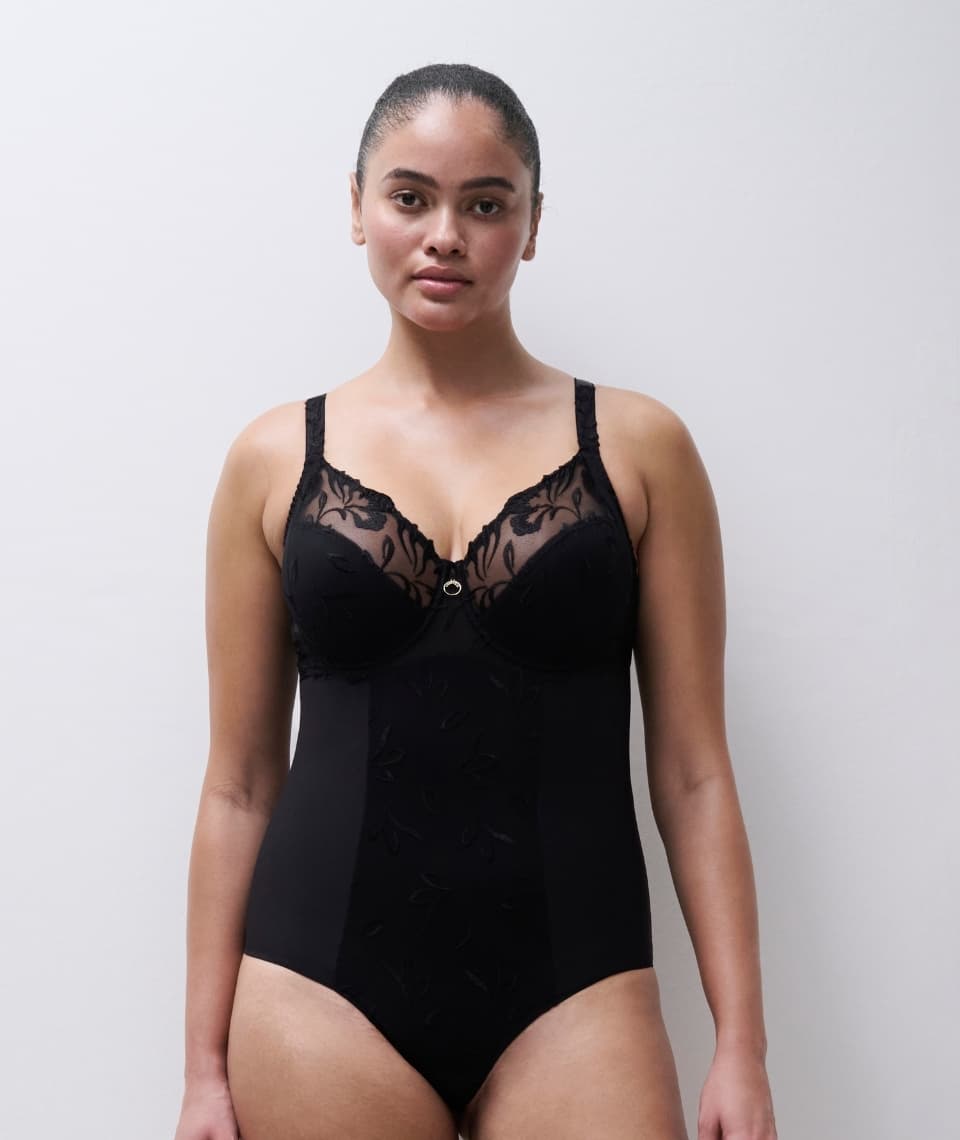 A person wearing a black lace bodysuit with floral patterns stands against a plain white background.
