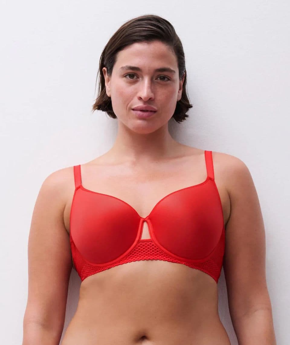 Person wearing a red bra with thin straps, standing against a plain white background, with short brown hair and a neutral expression.