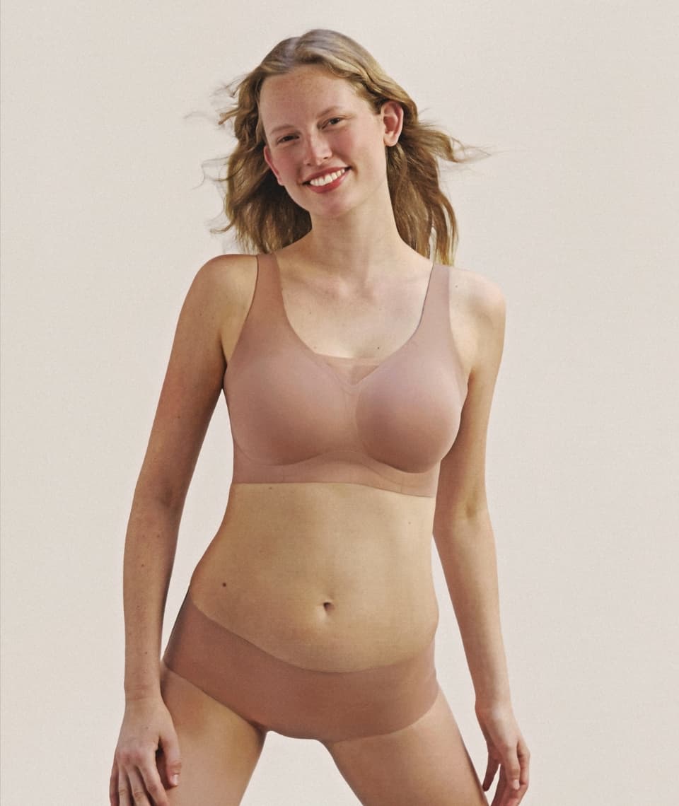 A smiling person with long hair poses in a seamless, nude-colored bra and underwear set against a plain background.