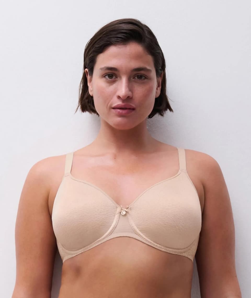 Person with short hair wearing a beige bra stands against a plain white background, gazing at the camera.