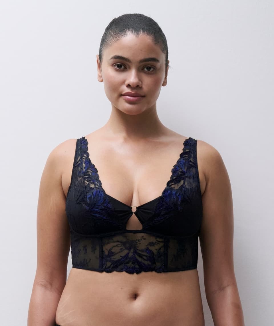 Woman wearing a black and blue lace bralette with floral patterns, standing against a plain white background.