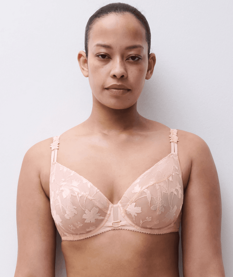 A person wearing a light pink lace bra with floral patterns, standing against a plain white background.