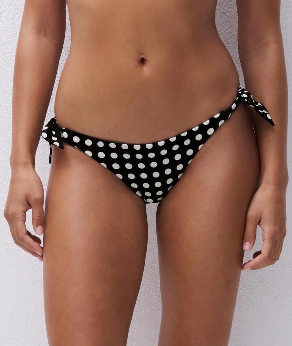 Person wearing black polka dot bikini bottoms with side ties, standing against a light wall.
