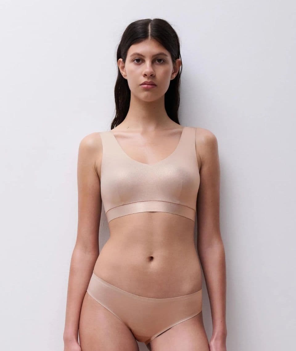 A woman stands against a plain background, wearing a matching nude-colored bra and panties.