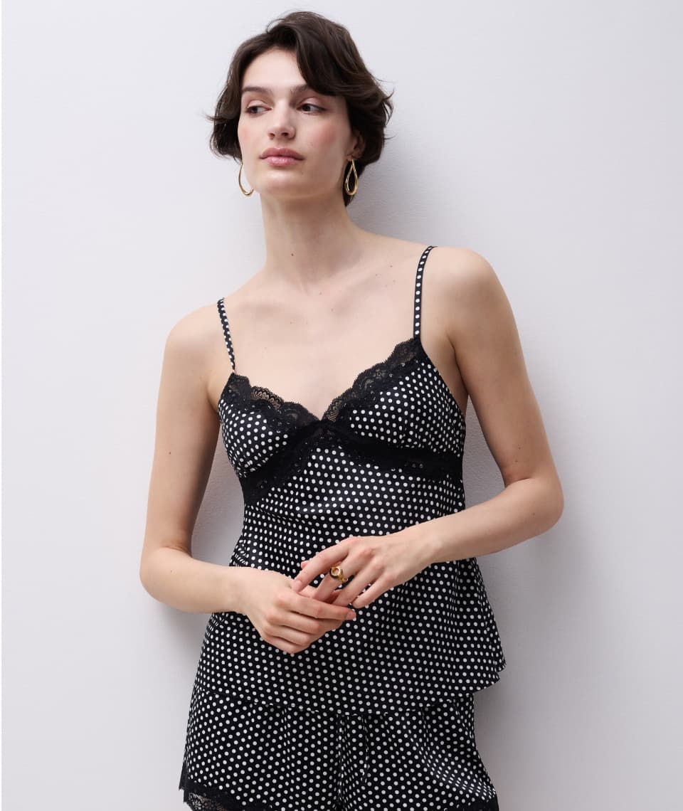 Person wearing a black polka dot camisole with lace trim, standing against a plain wall, looking slightly to the side.