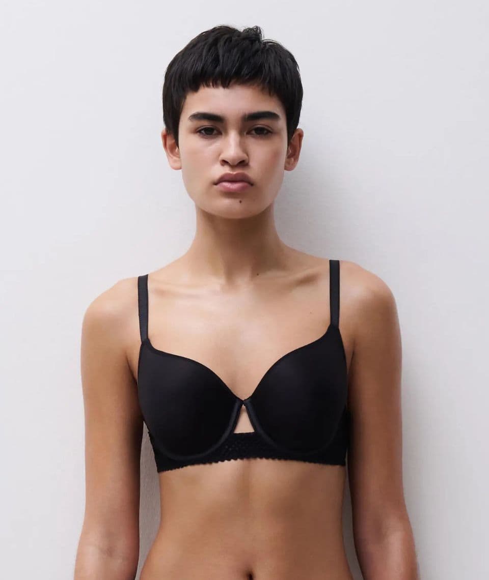 Person with short hair wearing a black bra stands against a plain white background, looking directly at the camera.