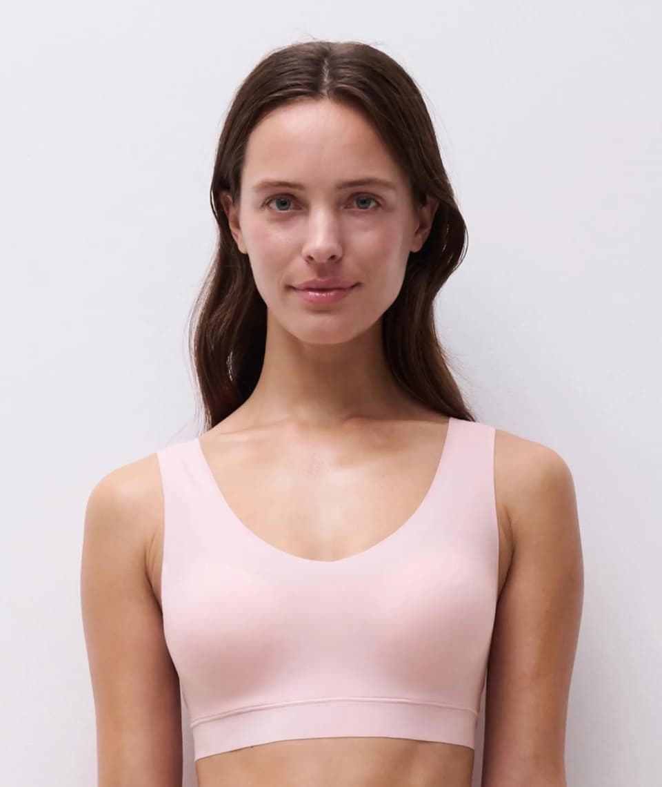 A woman with long brown hair wearing a light pink  bra, smiling slightly, against a plain white background.