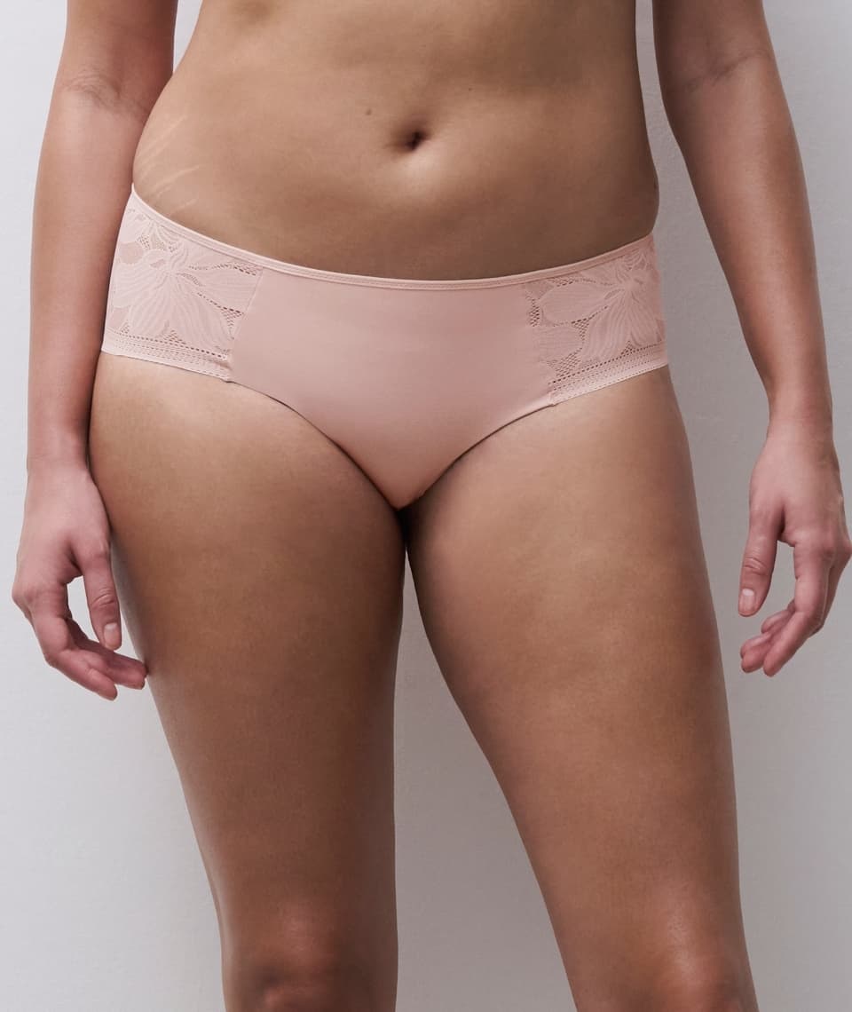 A person wearing pale pink underwear with floral lace detailing on the sides, standing against a plain backdrop.