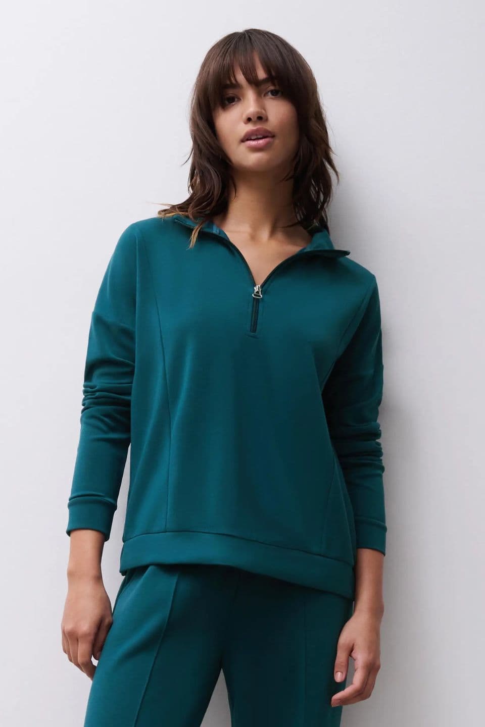 A woman is shown wearing a teal long-sleeve half-zip pullover with a relaxed fit.