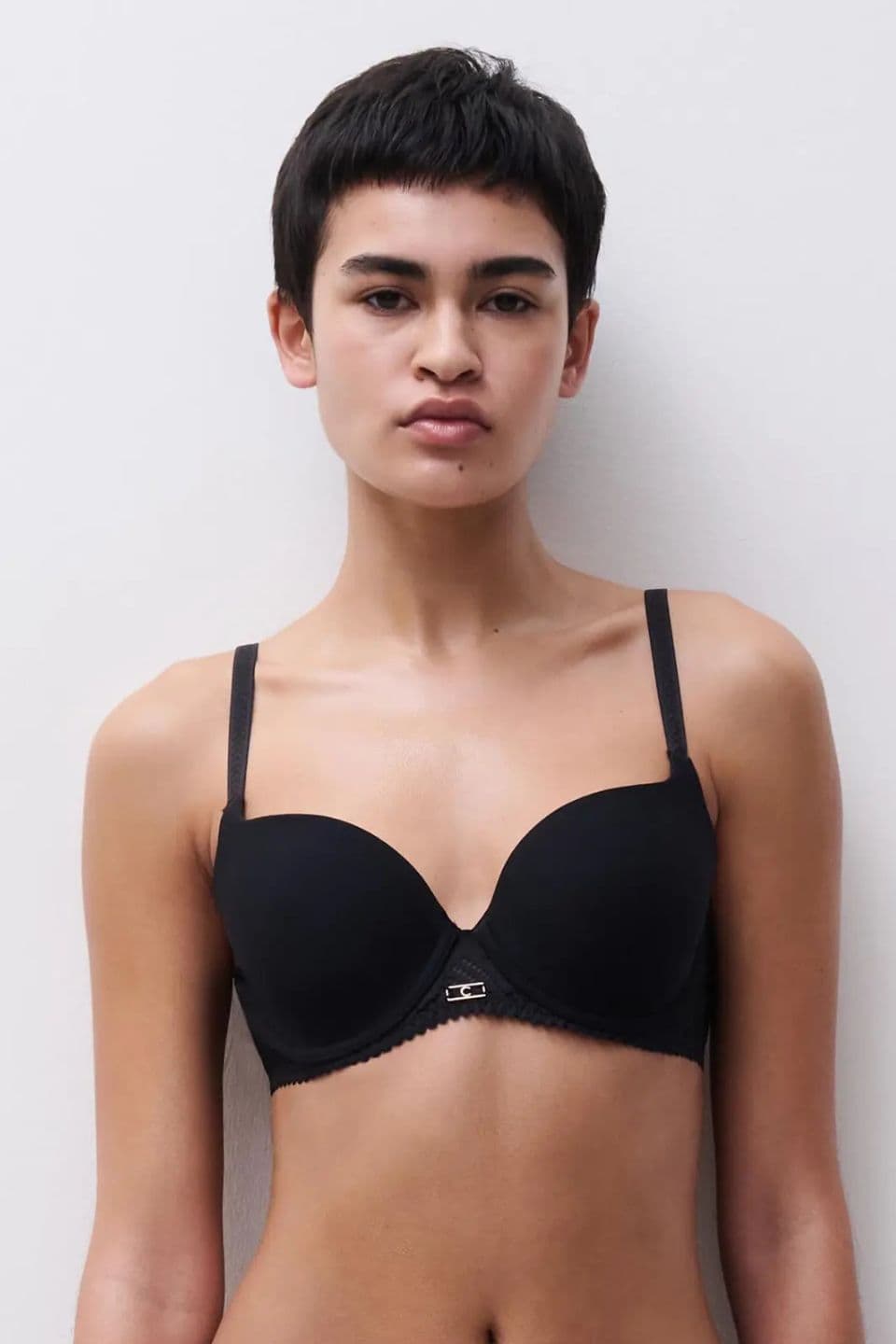 Person with short hair wearing a black bra against a plain background.