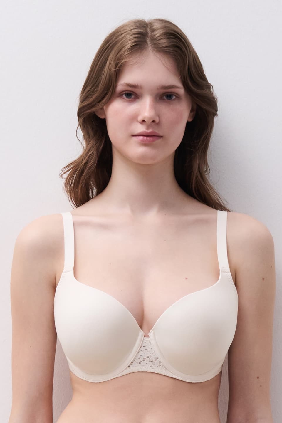 Woman wearing a cream-colored t-shirt bra with lace detailing, standing against a plain white background.