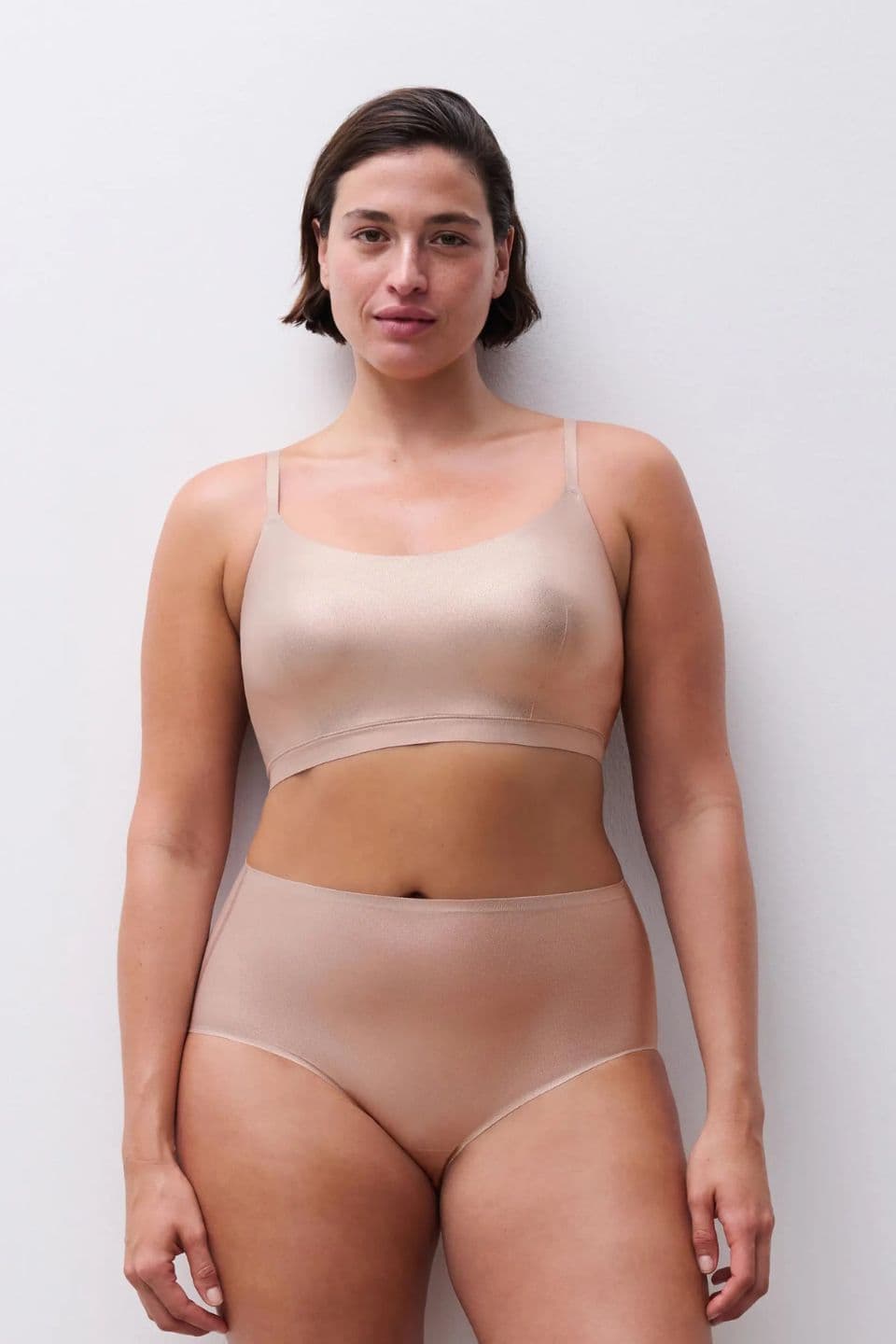 A woman wearing a nude-colored bra and panties set stands against a white background, with a confident and relaxed expression.