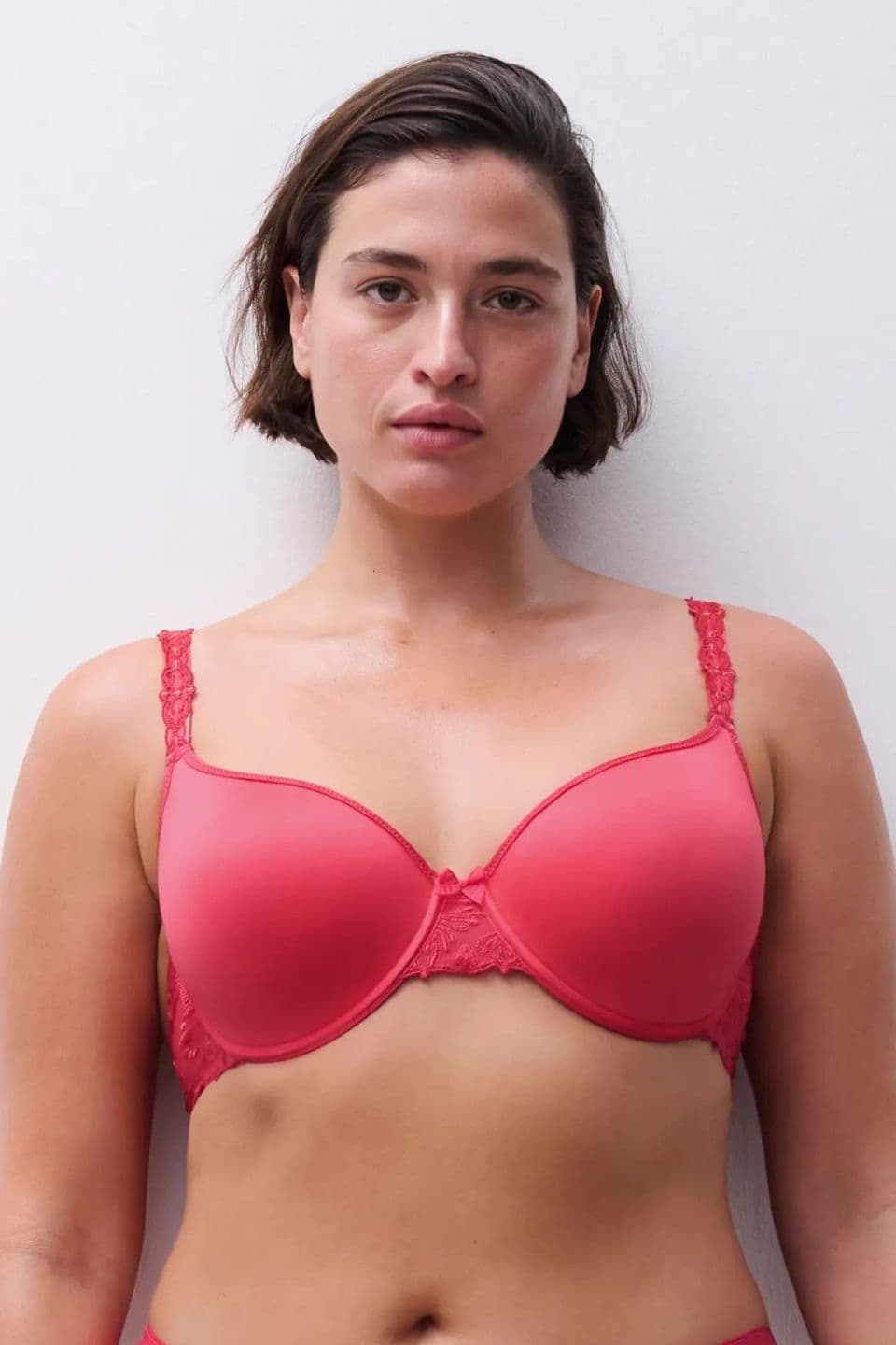 A person with short brown hair wearing a red bra stands against a plain white background, looking directly at the camera.