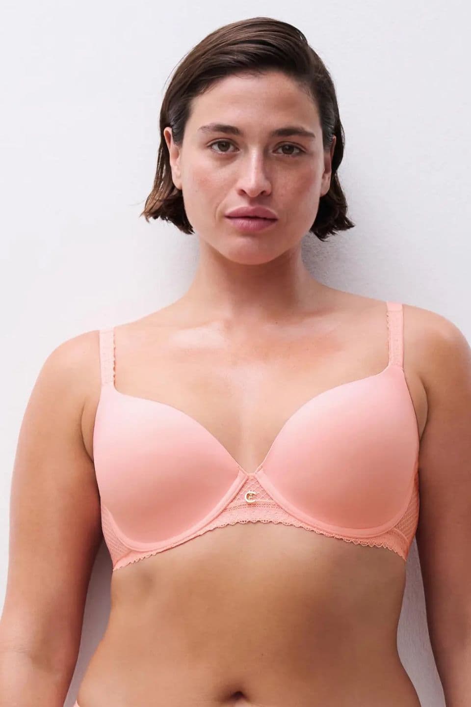 Woman in a peach-colored bra stands against a white background, looking directly at the camera, showcasing the lingerie.