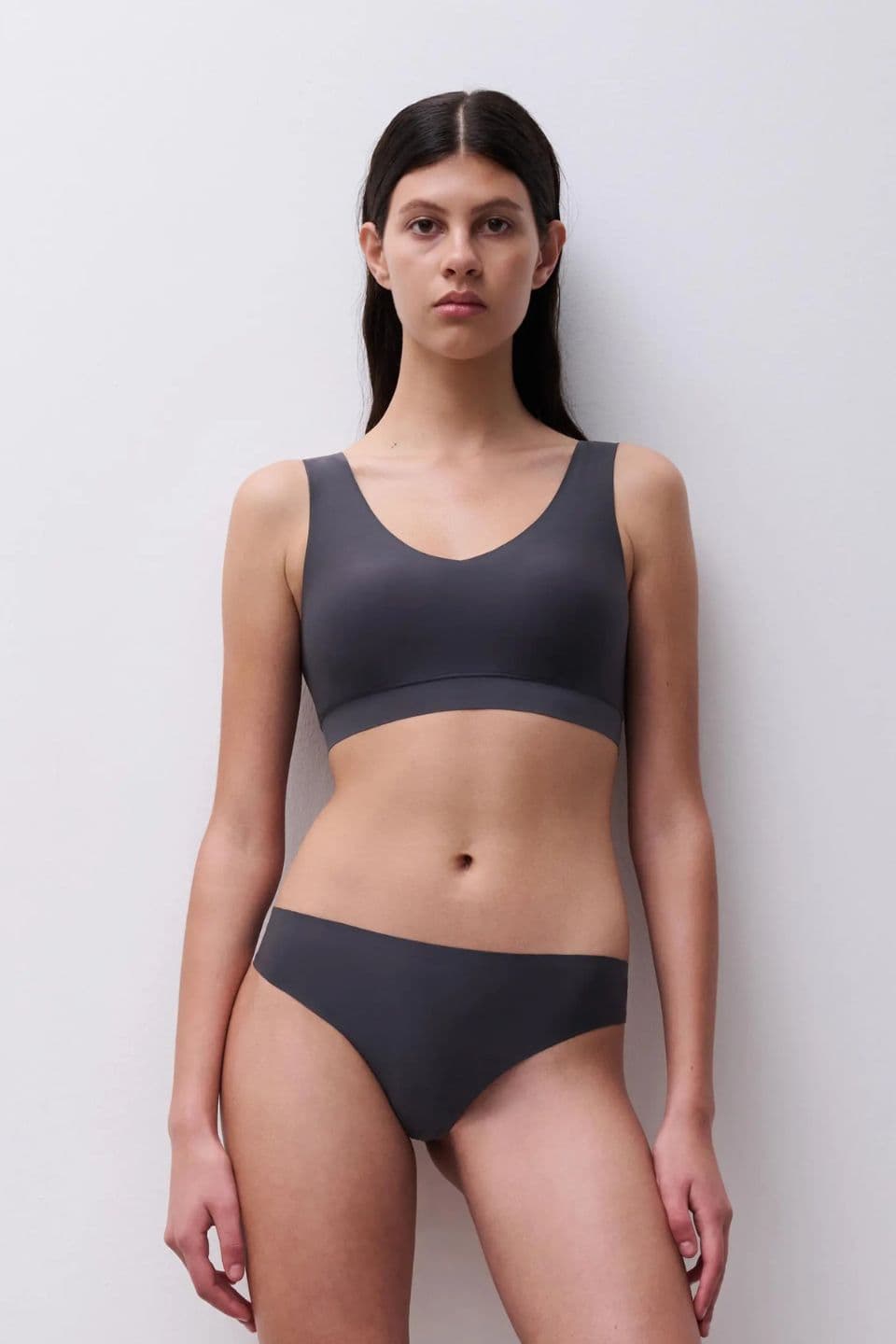 Woman wearing a matching dark gray bra and underwear, standing against a plain white wall.
