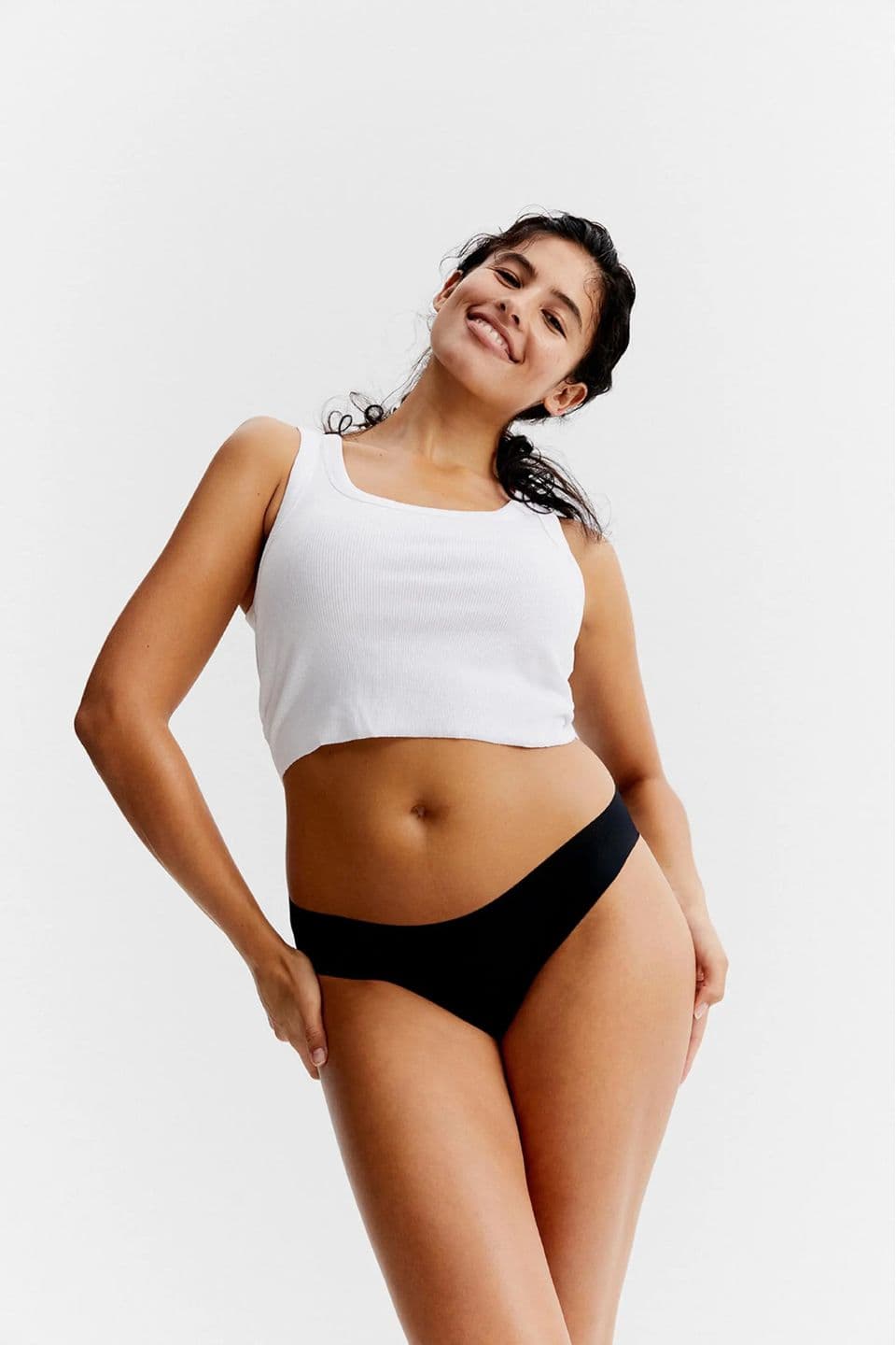 Smiling woman in a white cropped tank and black underwear posing against a white background, hands on hips.