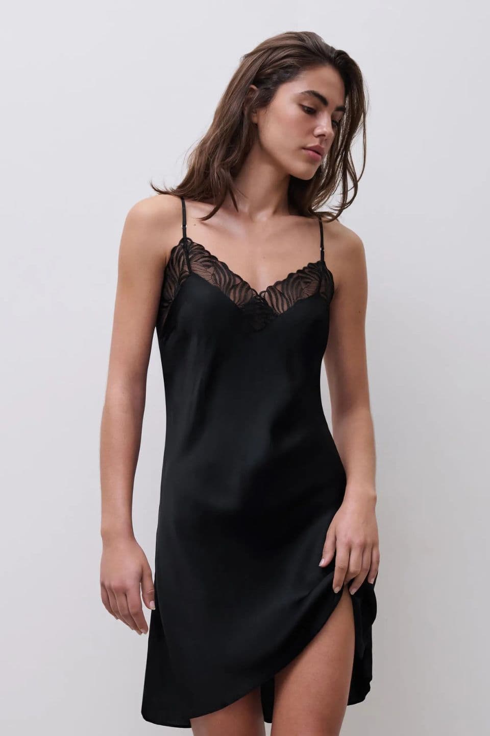 Woman wearing a black silk slip dress with lace trim and a thigh slit, looking down.