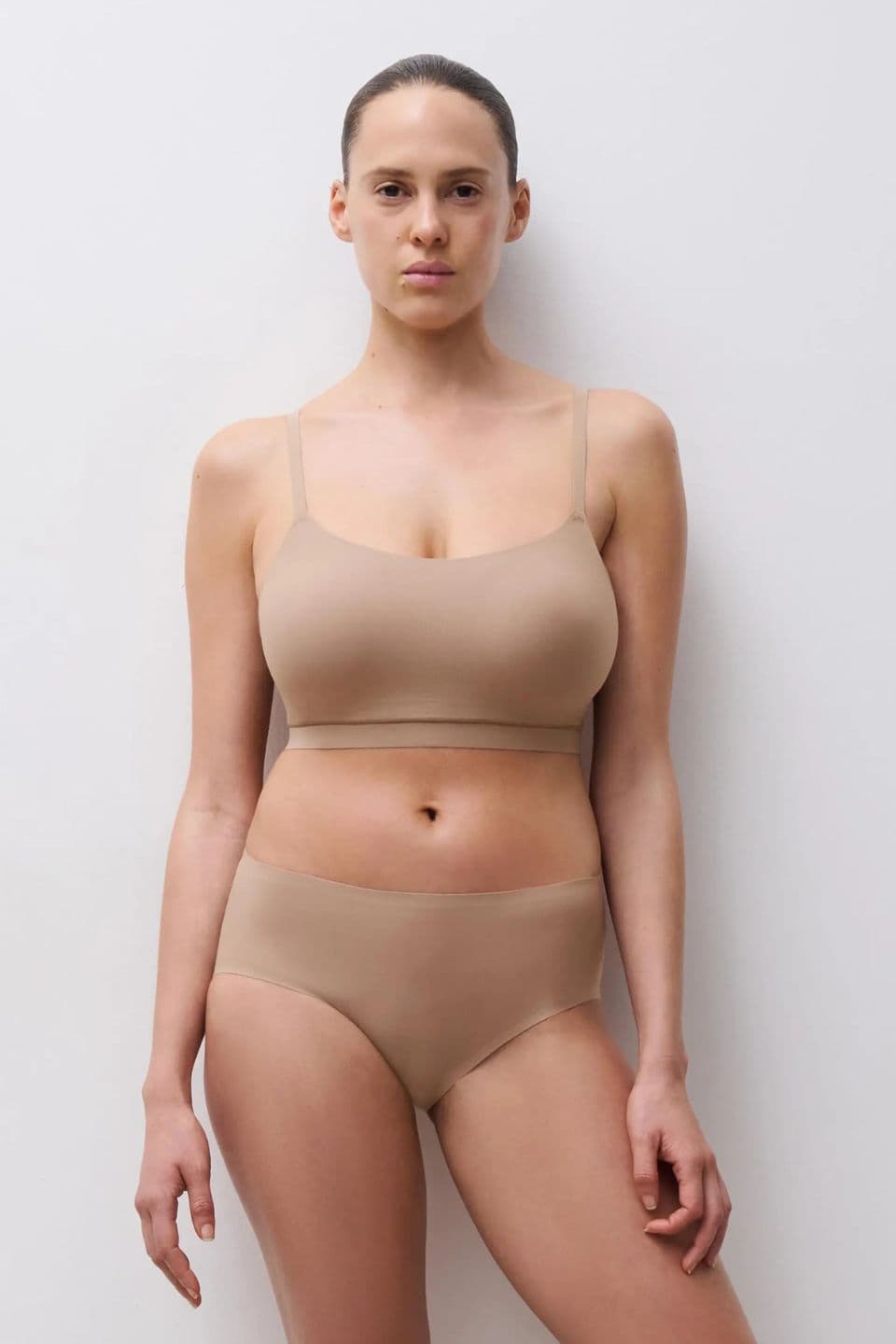 Person wearing a beige bralette and underwear, standing against a plain white background, with hair pulled back.